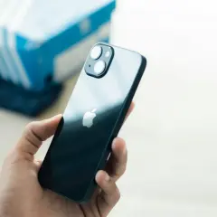 Dłoń trzyma ciemny iPhone 13. Czy iPhone 13 jest wodoszczelny? W tle niebieskie pudełko.