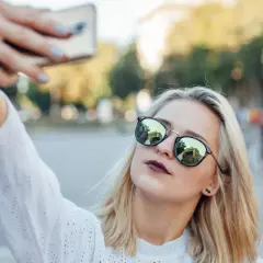 Blondynka w okularach robi sobie selfie telefonem. W okularach odbija się budynek. Oto jak zrobić dobre selfie telefonem.