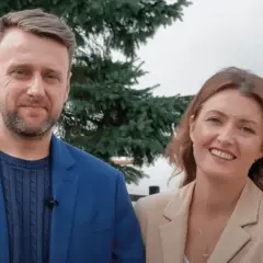 Uśmiechnięta para, on w niebieskiej marynarce, ona w beżowej. Wyglądają jak towarzystwo wzajemnej adoracji, ciesząc się wspólnym momentem.