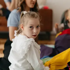Dziewczynka w białej bluzce patrzy w obiektyw, w tle kobieta i dziecko w wózku.