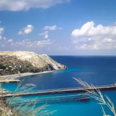 Panoramica delle isole Lipari con un porto e moli che si estendono nel mare blu intenso sotto un cielo azzurro con nuvole bianche.