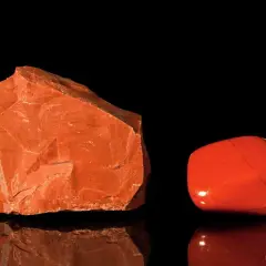 Zwei rote Jaspis-Arten: ein roher Stein und ein polierter Trommelstein, die auf schwarzem Hintergrund reflektieren.