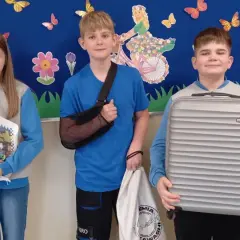 Pięcioro dzieci stoi przed ścianą z dekoracjami, trzymając walizki i torby zamiast plecaków.