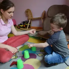 Terapia behawioralna autyzm ćwiczenia: chłopiec i terapeutka bawią się kubeczkami na dywanie.