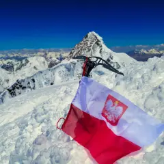 Na śnieżnym szczycie, z lodową siekierą i polską flagą, rozpościera się widok na skalisty, szeroki szczyt i góry.
