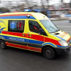 Żółto-czerwono-niebieska karetka Medicover z włączonymi sygnałami świetlnymi, w ruchu. Charakterystyczne oznaczenia karetek na karoserii.