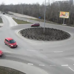 Widok z góry na rondo z kilkoma samochodami, drzewami i billboardami w tle.
