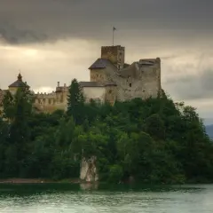Kamienny zamek na wzgórzu, otoczony zielenią, niczym z bajki o zamkach w okolicy Zakopanego. Woda lśni pod pochmurnym niebem.