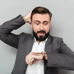 Hombre con traje gris, barba y expresión de pánico, se lleva una mano a la cabeza mientras mira su reloj. ¡Parece que va a soñar que llega tarde al trabajo!