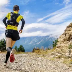 Un coureur en tenue de trail, sac sur le dos, s'élance sur un sentier rocailleux. Le paysage montagneux et le ciel bleu promettent une aventure ultra.
