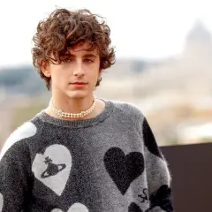 Timothée Chalamet con un ciuffo a tendina uomo, indossa un maglione grigio con cuori e una collana di perle.
