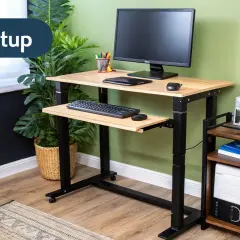 Adjustable height computer desk setup with monitor, keyboard, and mouse. A small side table holds a computer tower and books.