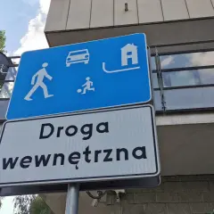 Znak "strefa zamieszkania" informuje o ograniczeniu prędkości. Widoczni piesi, dziecko grające w piłkę, samochód i dom.
