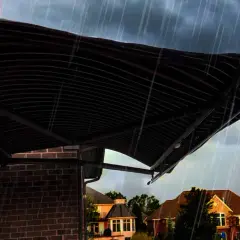 A dark, striped awning installation provides shelter from a heavy rain shower over a brick house.
