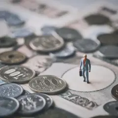 Mała figurka biznesmena stoi na tle japońskich banknotów i monet. To symboliczna wizja krajowej diety i finansów.
