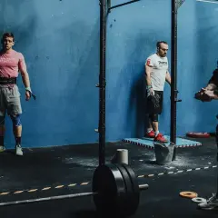 Trzech mężczyzn w siłowni CrossFit, jeden oparty o ścianę, drugi przy stojaku, trzeci z gumą treningową.