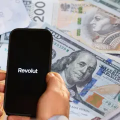 Telefon z logo Revolut na tle różnych walut. Może to symbolizować globalne finanse lub potencjalne zajęcie komornicze w kontekście międzynarodowych transakcji.