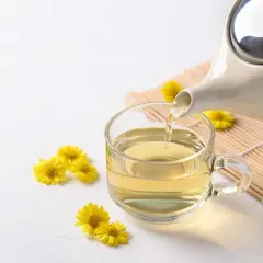 Tè giallo dorato versato da una teiera in una tazza trasparente, circondato da piccoli crisantemi fiori gialli.