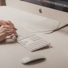 Mani che digitano su tastiera bianca, vicino a un mouse. Attenzione ai sintomi di tendinite da mouse per evitare dolori.
