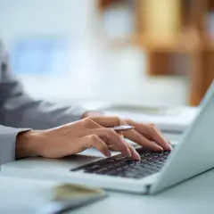Dłonie piszące na klawiaturze laptopa, w tle biuro.