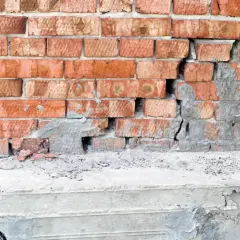 Uszkodzona ceglana ściana z widocznym pęknięciem w betonie, sugerującym podcinanie fundamentów.