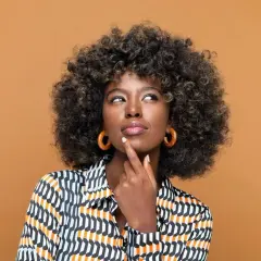 Mujer pensativa con cabello rizado y coloración semipermanente, luciendo una blusa estampada y aretes circulares.