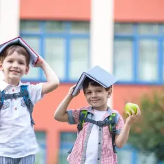 Dwóch chłopców z książkami na głowach i jabłkami w rękach przed szkołą. Zastanawiam się, czy to już 6 klasa, ile lat mają ci mali uczniowie?