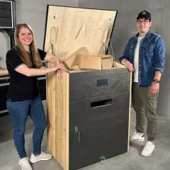 Ein Paar präsentiert stolz seine selbstgebaute Paketbox. Die Box ist aus Holz gefertigt und hat eine Klappe, die mit Ketten gesichert ist. Sie ist groß genug, um mehrere Pakete aufzunehmen.