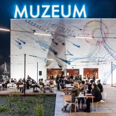 Wieczorem muzeum sztuki nowoczesnej nad Wisłą tętni życiem. Ludzie siedzą przy stolikach na zewnątrz, podziwiając abstrakcyjną grafikę na ścianie budynku.