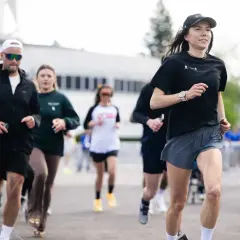 Grupa ludzi biegnie, w tym kobieta w czarnej koszulce i spodenkach, która może być inspiracją, jak polubić bieganie. W tle widać osoby na wózkach.