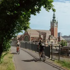 Dwie osoby na rowerach jadą ścieżką obok torów kolejowych. W tle piękny budynek dworca z wieżą zegarową. To idealne miejsce przyjazne rowerzystom.