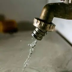 Un robinet en laiton coule, projetant un jet d'eau. Des éponges floues sont visibles en arrière-plan. Il est temps d'avoir plus d'eau dans la maison.