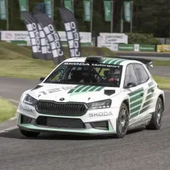 Biała Skoda Fabia Rally z zielonymi pasami pędzi po torze. Kierowca w kasku jest gotowy do akcji.