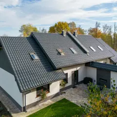 Nowoczesny dom z białymi ścianami i ciemnym, eleganckim dachem z dachówki ceramicznej. Wokół zieleń i jesienne drzewa.