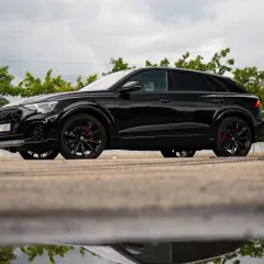 Czarny Audi Q9 z czerwonymi zaciskami hamulcowymi, odbijający się w kałuży. Sprawdź audi q9 cena!