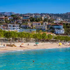 Piękna, słoneczna **Albania** w **czerwcu**. Ludzie odpoczywają na plaży, woda jest turkusowa, a miasteczko malowniczo wznosi się na wzgórzu.