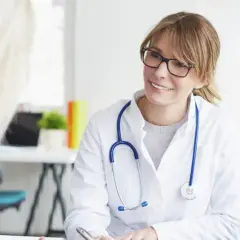 Lekarka w białym kitlu z niebieskim stetoskopem uśmiecha się do pacjentki. Rozmowa o zdrowiu, być może próba tężyczkowa.