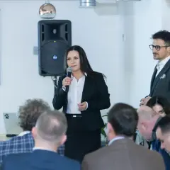 Kobieta w czarnej marynarce mówi do mikrofonu, przed nią siedzą ludzie.