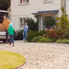 Rodzina rozpakowuje auto na podjeździe. Jaki kliniec na podjazd, chłopiec z deskorolką biegnie do domu, gdzie czeka mama z córką.
