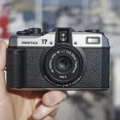 A hand holds the highest quality film camera, a silver and black Pentax 17, with a blurred marina in the background.