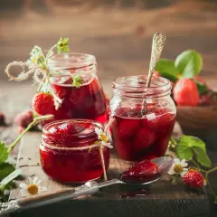 Słoiki z domowymi truskawkami w syropie, ozdobione sznurkiem i kwiatkami. Obok świeże owoce i łyżeczka z konfiturą.