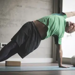 Hombre mayor con barba gris practica yoga para hombres en una esterilla, en postura de plancha lateral.