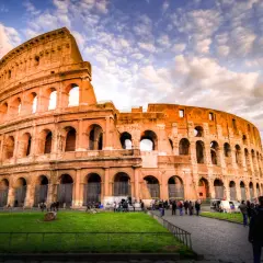 Il Colosseo, un'icona di cosa visitare a Roma, si erge maestoso sotto un cielo drammatico. Turisti passeggiano sul selciato, ammirando l'antica grandezza.