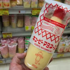 A hand holds a large squeeze bottle of Kewpie mayonnaise. The packaging features the iconic Kewpie doll. The question "how long does Kewpie mayo last?" is on my mind as I shop.