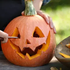 Händer skär ett ansikte i en pumpa för en klassisk pumpasminkning. Bredvid ligger en skål med pumpafrön och fruktkött.