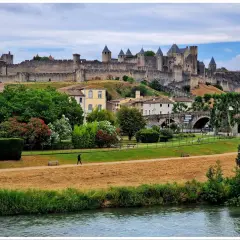 Majestatyczna twierdza Carcassonne góruje nad miastem. W dole zielony park, rzeka i spacerująca postać.