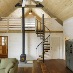Idee soppalco basso: scala a chiocciola in metallo e legno porta a un soppalco con ringhiera in legno, stufa a legna e divano.
