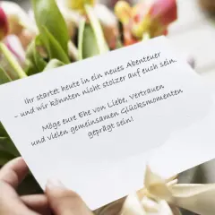 Hände halten eine Karte mit Hochzeitsglückwünschen vor Tulpen. "Ihr startet heute in ein neues Abenteuer – und wir könnten nicht stolzer auf euch sein. Möge eure Ehe von Liebe, Vertrauen und vielen gemeinsamen Glücksmomenten geprägt sein!