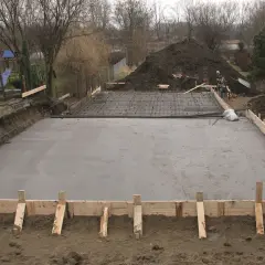 Fundamenty pod dom: wylana płyta betonowa z zbrojeniem, szalunki drewniane, koparka w tle.