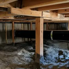 Synlig fukt i krypgrund. En stor svart rörledning löper genom utrymmet under huset, där det syns jord och byggmaterial.